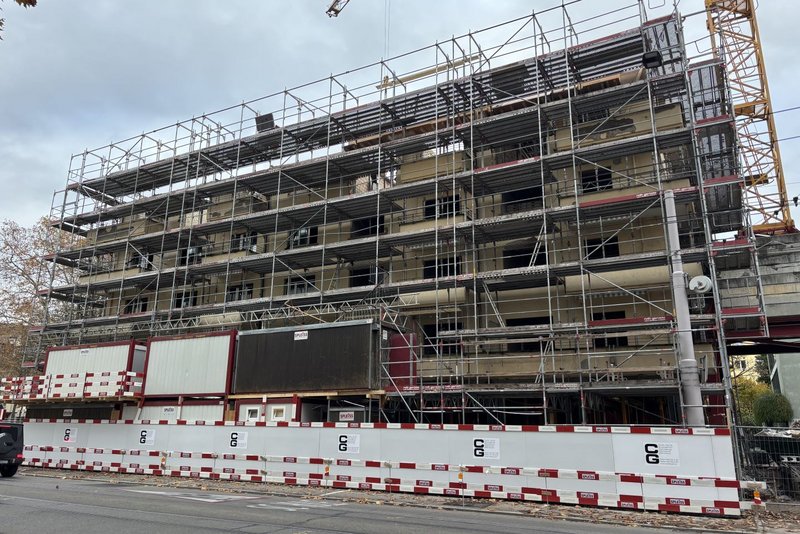 Frontansicht eines mehrstöckigen Gebäudes in der Bauphase: Ein grosses Baugerüst umschliesst die Fassade, unten sind Baustellenabschrankungen und Container zu sehen; die Fassade ist noch offen und wird saniert. Das Bild zeigt die Ausgangssituation für eine Gerüstverkleidung bzw. Baugerüst-Gestaltung im Grossformat, wie sie Richnerstutz für Kunst am Bau umsetzt.