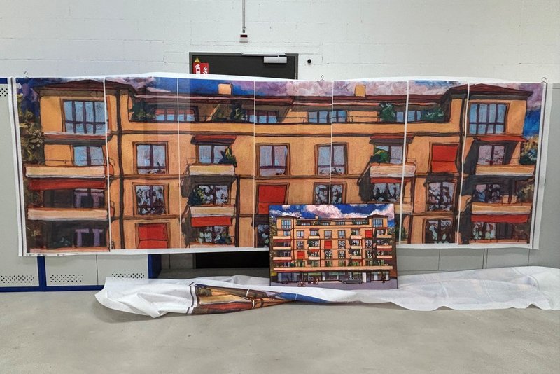 In einer Werkstatt hängt ein breites, segmentiertes Banner mit einer gemalten Hausfassade in warmen Orange- und Rottönen; davor steht das kleinere Originalbild bzw. eine Vorlage auf einer Staffelei, unten liegt weiteres bedrucktes Material. Das Foto zeigt die Übertragung eines Acrylgemäldes von Michele Bordoni in den Grossformatdruck für Fassadenkunst, also die Druckabstimmung für den Blachendruck der späteren Gerüstverkleidung als Baugerüst-Gestaltung, umgesetzt von Richnerstutz. 