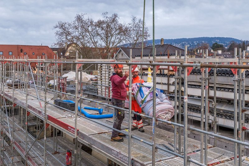 Zwei Arbeiter stehen auf einem Baugerüst und führen mit einem Kran eine grosse, aufgerollte Blache/Gitternetz-Blache an Gurten heran; im Hintergrund sind Bahngleise und Gebäude zu sehen. Die Szene zeigt die Vorbereitung der Gerüstverkleidung bzw. Baugerüst-Gestaltung im Grossformat – ein Schritt der Montage für Kunst am Bau, umgesetzt von Richnerstutz als Grossformatdruck bzw. Blachen-Druck.