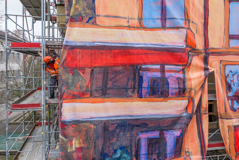 Nahaufnahme eines Baugerüsts: Ein Monteur in Sicherheitsausrüstung arbeitet seitlich an einer grossen, bedruckten Gitternetz-Blache, die das Gerüst verhüllt. Das Motiv zeigt in kräftigen Orange-, Rot- und Blautönen die hochauflösende Grossformat-Reproduktion einer gemalten Hausfassade, umgesetzt als Grossformatdruck für die Gerüstverkleidung durch Richnerstutz.