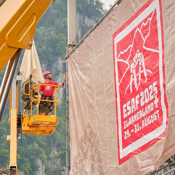 Monteure bringen auf einer Hebebühne eine grosse Blache mit dem ESAF-2025-Design an einer Tribüne an.
