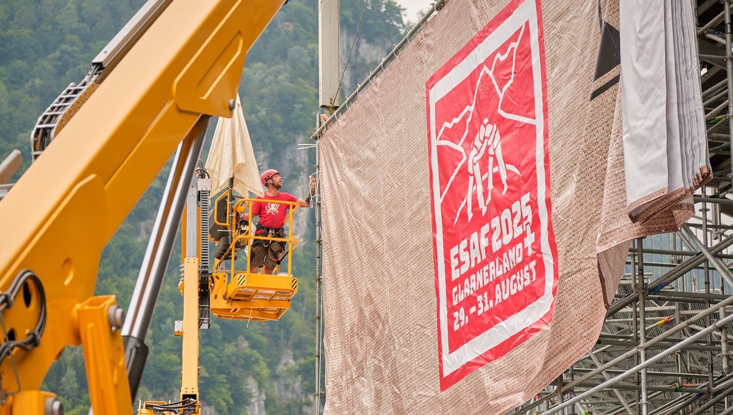 Monteure bringen auf einer Hebebühne eine grosse Blache mit dem ESAF-2025-Design an einer Tribüne an.