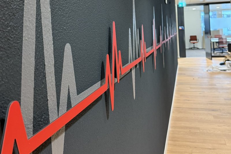 Rote Herzschlag-Grafik aus Dibond mit 3D-Abstandshaltern vor grauer Folienkontur.
