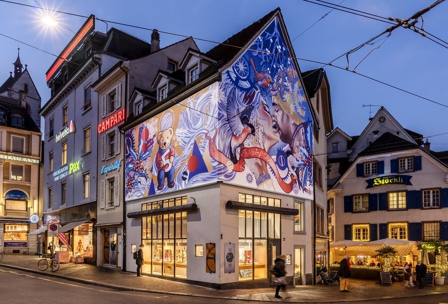 Dämmerungsaufnahme in Basel: Das Eckgebäude des Maison Oris trägt eine temporäre Fassadenverkleidung mit grossformatigen, farbigen Illustrationen von Patrizia Stalder und setzt zur Adventszeit einen auffälligen weihnachtlichen Akzent.