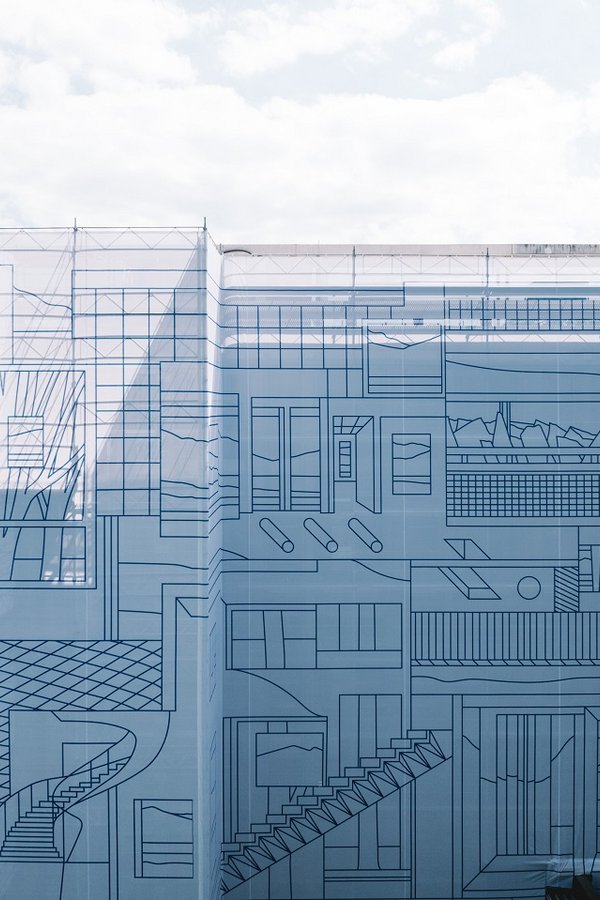 Frontalansicht eines Gebäudes, das vollständig mit einer hellen Gerüstverkleidung eingekleidet ist; auf der riesigen Plane sind feine, blaue Linien als architektonische Zeichnung über die ganze Fassade gedruckt. Das Bild zeigt die Grossformat-Umsetzung und Baugerüst-Einkleidung für den Hauptsitz der Vaudoise Versicherungen in Lausanne – das Kunstprojekt «On and Around» von Nigel Peake, umgesetzt von Richnerstutz.