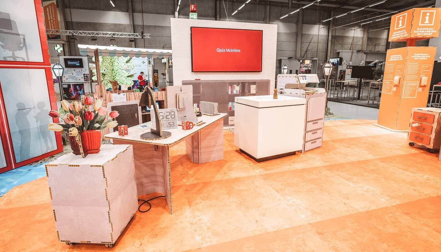 SwissSkills exhibition stand made from honeycomb cardboard. Die Mobiliar created a 300 m² exhibition stand from this material. Except for a few items such as screens, all elements and accessories are made of cardboard, including: bouquet, table lamp, desk, clock, chest of drawers, partition wall with integrated screen, sales counter, etc.