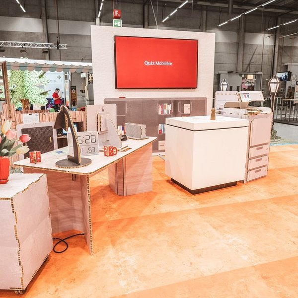 Stand SwissSkills en carton alvéolé. Die Mobiliar a réalisé un stand de 300 m² avec ce matériau. À l’exception de quelques éléments comme les écrans, tous les éléments et accessoires sont en carton : bouquet de fleurs, lampe de table, bureau, horloge, commode, cloison avec écran intégré, comptoir de vente, etc.