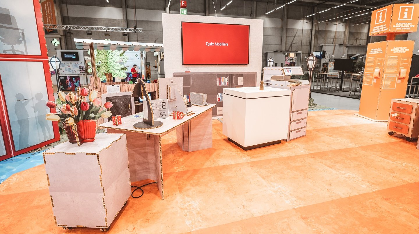 SwissSkills Messestand aus Wabenkarton. Die Mobiliar hat 300 m2 Messestand aus diesem Mobiliar gefertigt. Bis auf ein paar Ausnahmen wie Screens sind alle Elemente und Accessoires aus Karton, wie: Blumenstrauss, Tischlampe, Schreibtisch, Uhr, Kommode, Stellwand mit integriertem Screen, Verkaufsstand usw.