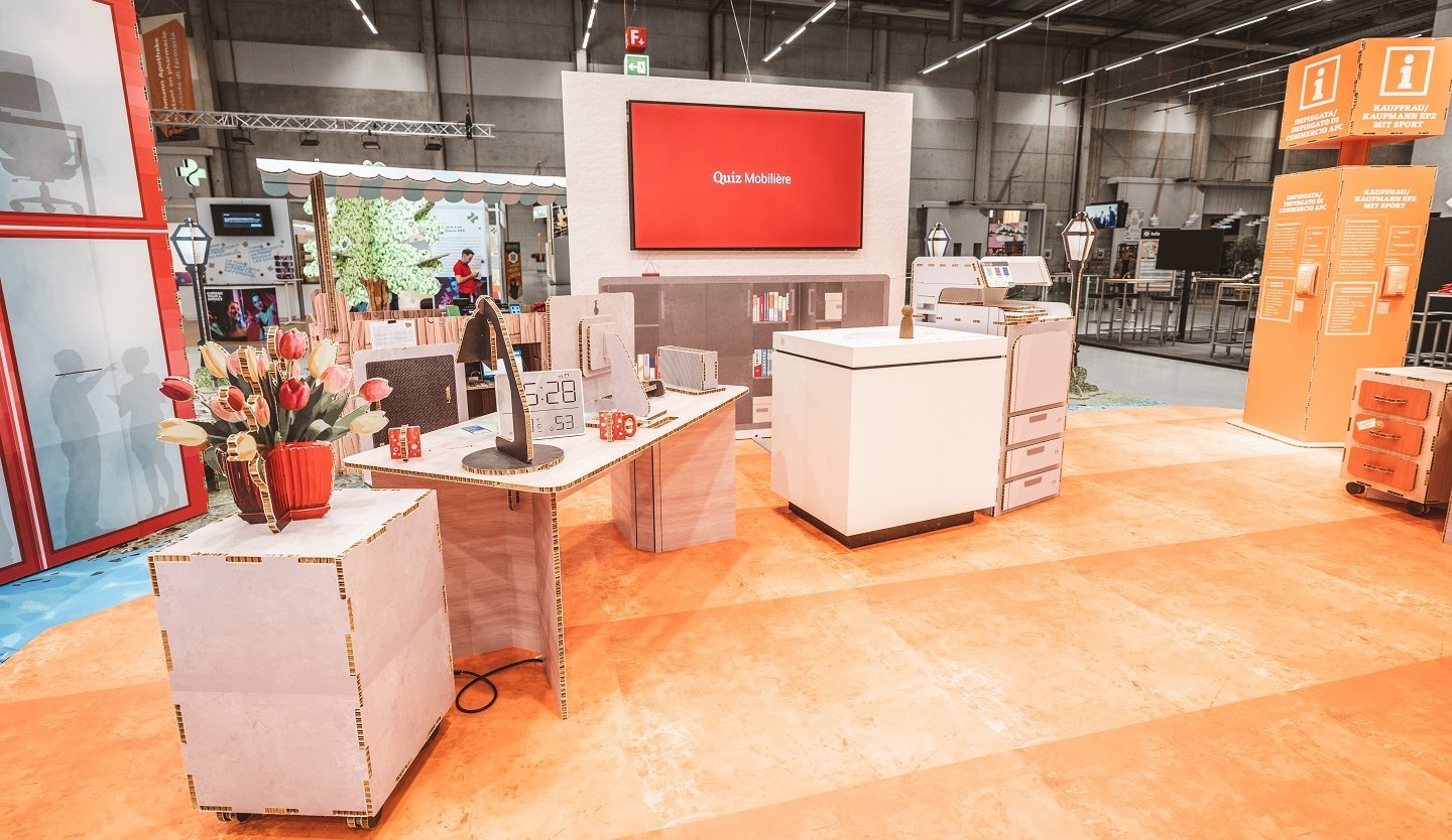 Stand SwissSkills en carton alvéolé. Die Mobiliar a réalisé un stand de 300 m² avec ce matériau. À l’exception de quelques éléments comme les écrans, tous les éléments et accessoires sont en carton : bouquet de fleurs, lampe de table, bureau, horloge, commode, cloison avec écran intégré, comptoir de vente, etc.