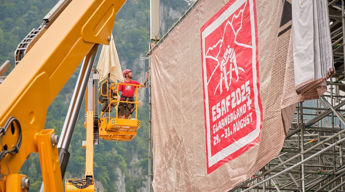Alternative text    Fitters use a lifting platform to attach a large tarpaulin with the ESAF 2025 design to a grandstand.