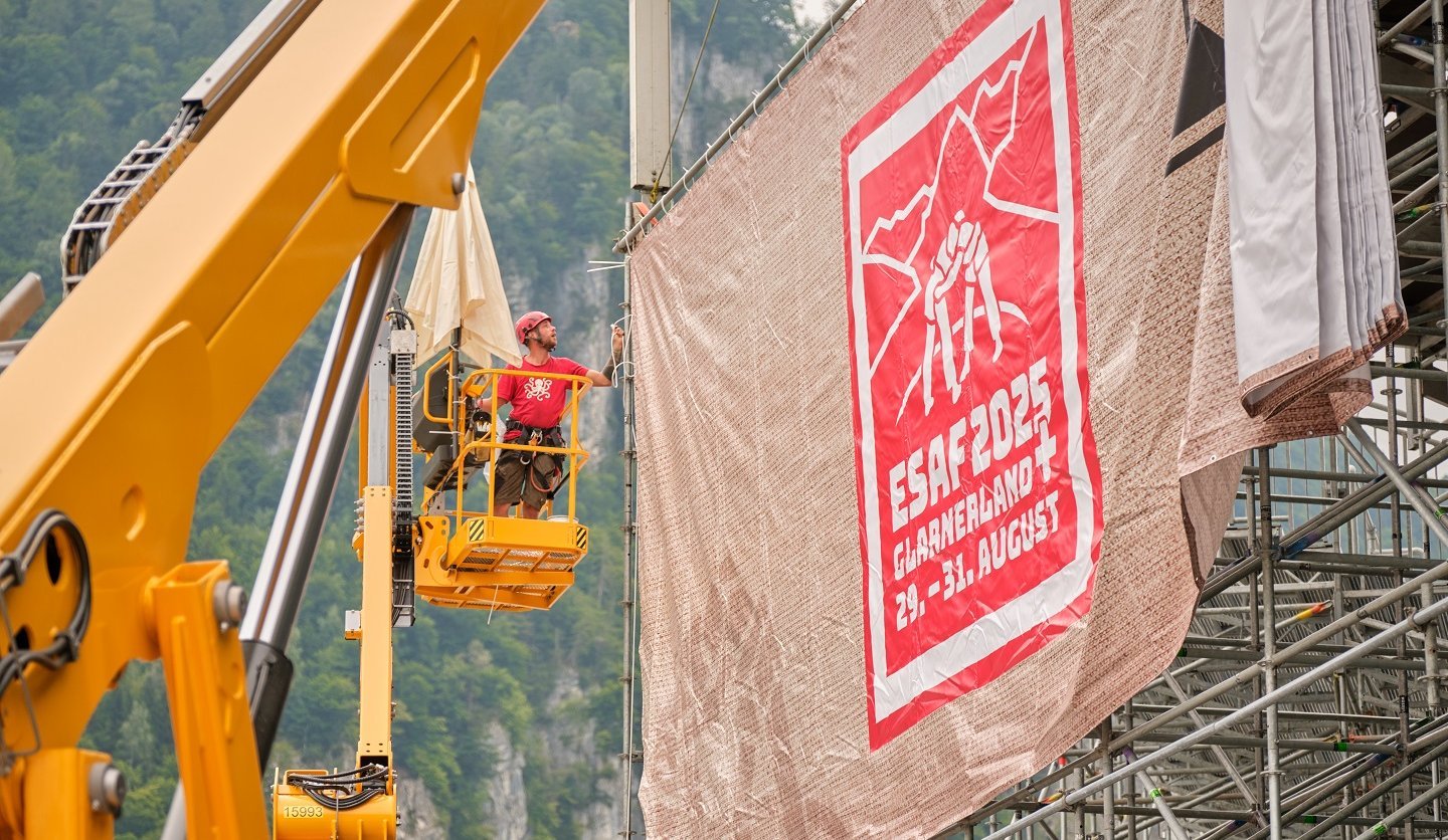 Des monteurs installent sur une nacelle une grande bâche avec le design ESAF 2025 sur un gradin.