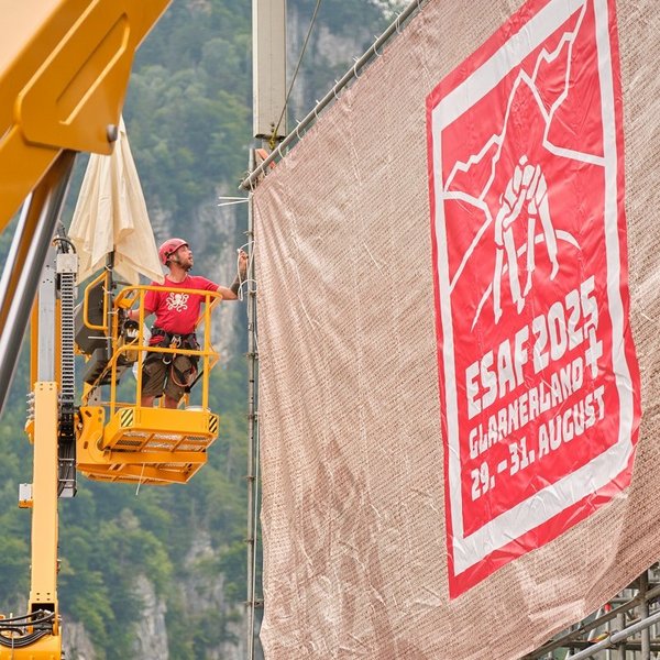 Alternative text    Fitters use a lifting platform to attach a large tarpaulin with the ESAF 2025 design to a grandstand.