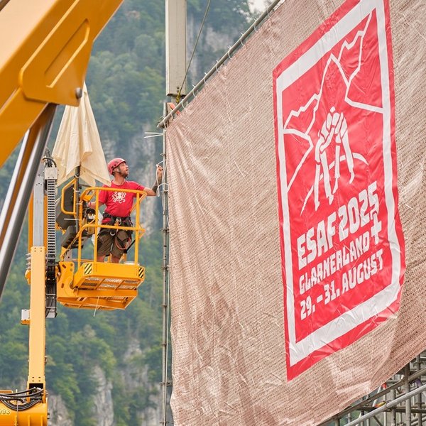 Des monteurs installent sur une nacelle une grande bâche avec le design ESAF 2025 sur un gradin.