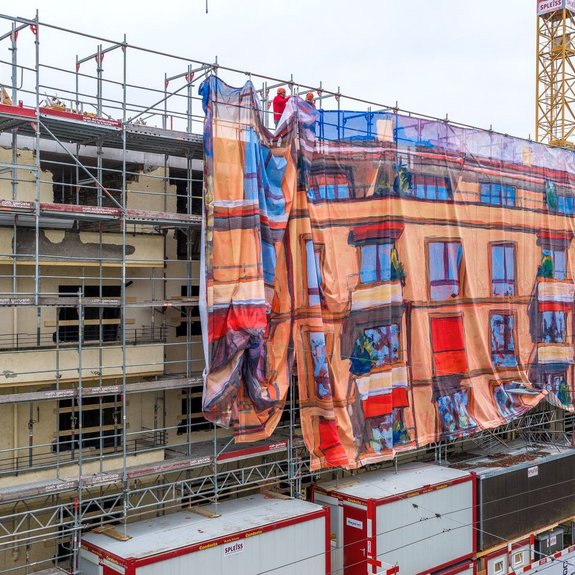 Grossaufnahme eines eingerüsteten Mehrfamilienhauses: Ein grosses Baugerüst wird teilweise mit einer bedruckten Gitternetz-Blache verhüllt, die wie Fassadenkunst wirkt und die Hausfassade als farbiges Bildmotiv nachzeichnet; oben befestigen Monteure die Gerüstverkleidung. Die Baugerüst-Gestaltung im Grossformat wurde von Richnerstutz als Grossformatdruck umgesetzt – basierend auf einem Acrylgemälde von Michele Bordoni, hochauflösend reproduziert durch undknup AG, im Auftrag von Urs Ledermann.