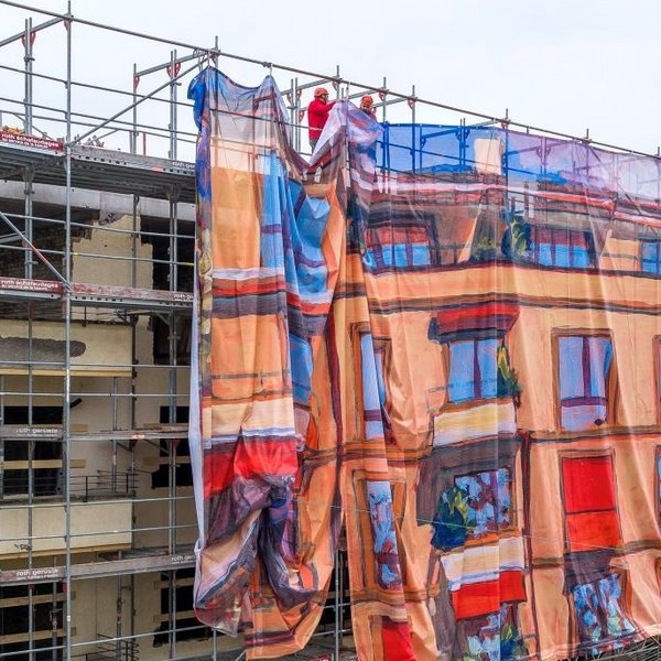 Grossaufnahme eines eingerüsteten Mehrfamilienhauses: Ein grosses Baugerüst wird teilweise mit einer bedruckten Gitternetz-Blache verhüllt, die wie Fassadenkunst wirkt und die Hausfassade als farbiges Bildmotiv nachzeichnet; oben befestigen Monteure die Gerüstverkleidung. Die Baugerüst-Gestaltung im Grossformat wurde von Richnerstutz als Grossformatdruck umgesetzt – basierend auf einem Acrylgemälde von Michele Bordoni, hochauflösend reproduziert durch undknup AG, im Auftrag von Urs Ledermann.