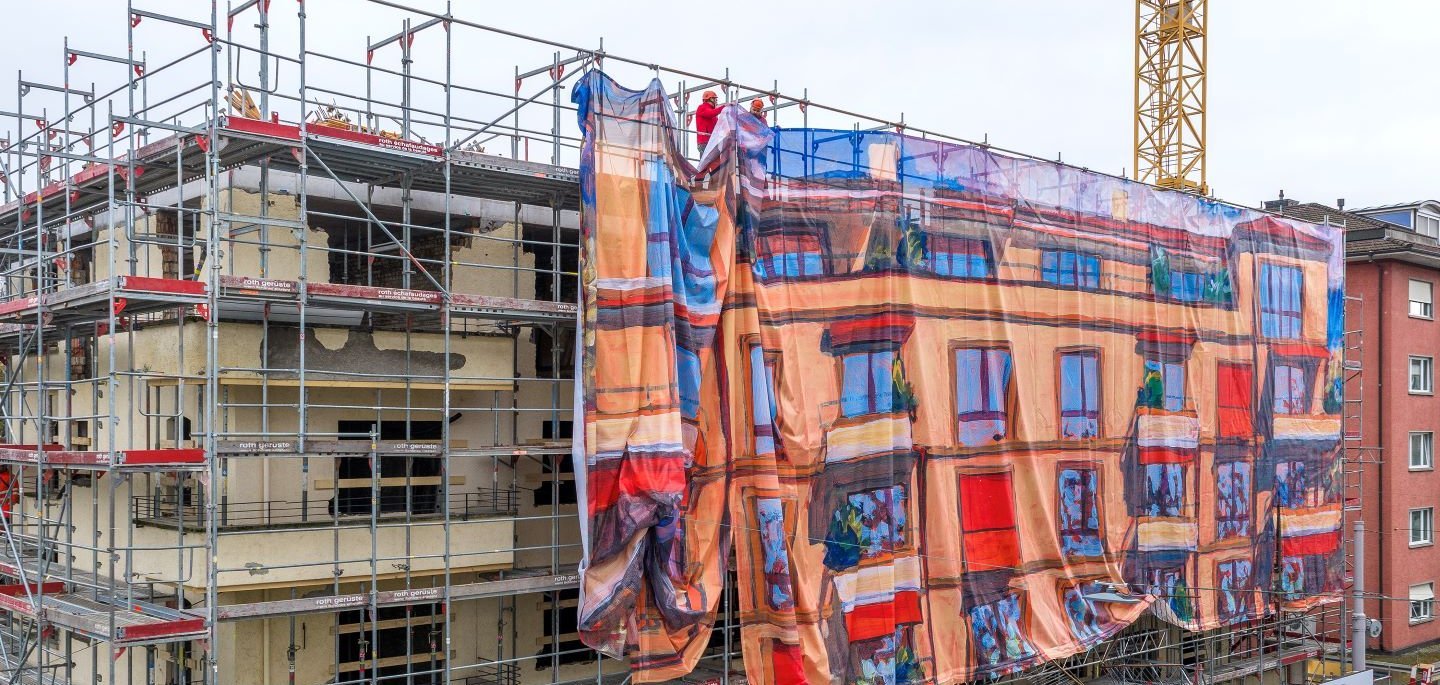 Grossaufnahme eines eingerüsteten Mehrfamilienhauses: Ein grosses Baugerüst wird teilweise mit einer bedruckten Gitternetz-Blache verhüllt, die wie Fassadenkunst wirkt und die Hausfassade als farbiges Bildmotiv nachzeichnet; oben befestigen Monteure die Gerüstverkleidung. Die Baugerüst-Gestaltung im Grossformat wurde von Richnerstutz als Grossformatdruck umgesetzt – basierend auf einem Acrylgemälde von Michele Bordoni, hochauflösend reproduziert durch undknup AG, im Auftrag von Urs Ledermann.