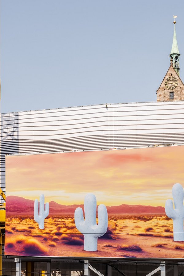 Detailaufnahme Grossformatige Kunstinstallation von Claudia Comte am historischen Globus-Gebäude auf dem Marktplatz Basel: Im Vordergrund ist eine bedruckte Blache mit einer Wüstenlandschaft und mehreren stilisierten, hellen Kakteen vor einem warmen, orange-gelben Himmel zu sehen. Direkt darüber erkennt man ein Gitternetz mit schwarz-weissen, wellenartigen Linienmustern, das die drunterliegende Gebäudefassade umhüllt.