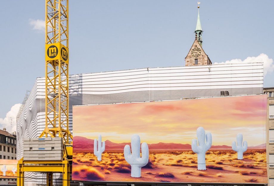 Detailaufnahme Grossformatige Kunstinstallation von Claudia Comte am historischen Globus-Gebäude auf dem Marktplatz Basel: Im Vordergrund ist eine bedruckte Blache mit einer Wüstenlandschaft und mehreren stilisierten, hellen Kakteen vor einem warmen, orange-gelben Himmel zu sehen. Direkt darüber erkennt man ein Gitternetz mit schwarz-weissen, wellenartigen Linienmustern, das die drunterliegende Gebäudefassade umhüllt.