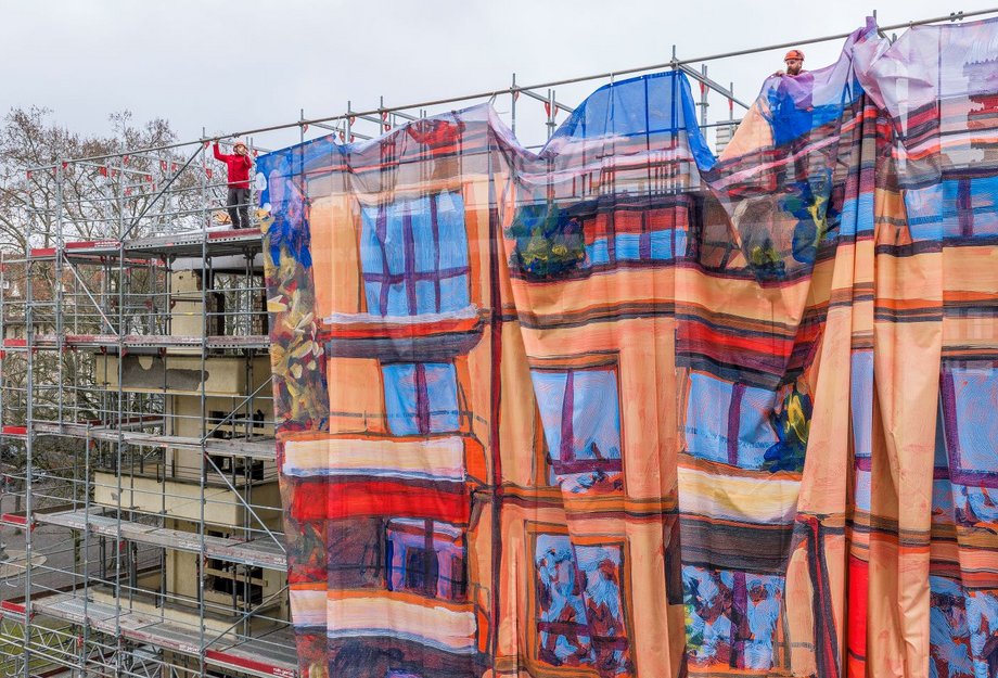 Ein mehrstöckiges Gebäude ist eingerüstet; mehrere Monteure befestigen oben eine grosse, farbig bedruckte Gitternetz-Blache am Baugerüst. Das Motiv zeigt eine gemalte Hausfassade und wirkt als Kunst am Bau – eine Grossformat-Reproduktion eines Acrylgemäldes von Michele Bordoni, umgesetzt als Grossformatdruck für die Gerüstverkleidung durch Richnerstutz.