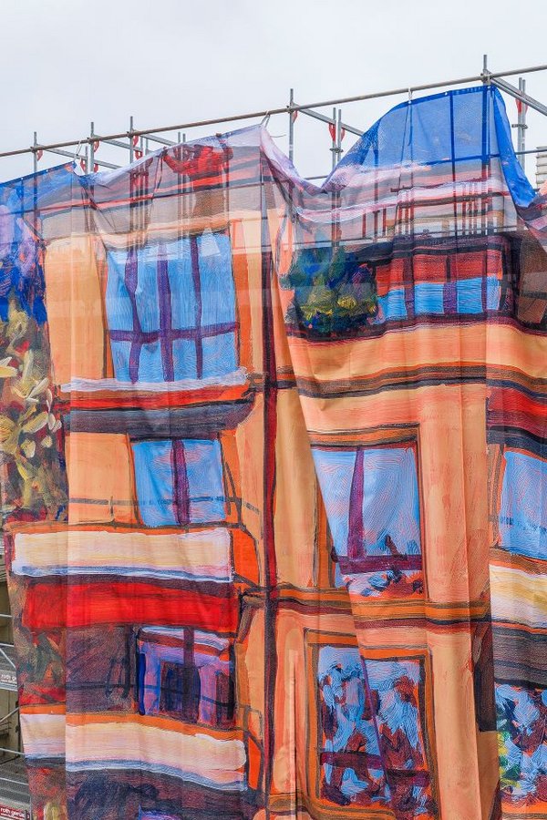 Ein mehrstöckiges Gebäude ist eingerüstet; mehrere Monteure befestigen oben eine grosse, farbig bedruckte Gitternetz-Blache am Baugerüst. Das Motiv zeigt eine gemalte Hausfassade und wirkt als Kunst am Bau – eine Grossformat-Reproduktion eines Acrylgemäldes von Michele Bordoni, umgesetzt als Grossformatdruck für die Gerüstverkleidung durch Richnerstutz.