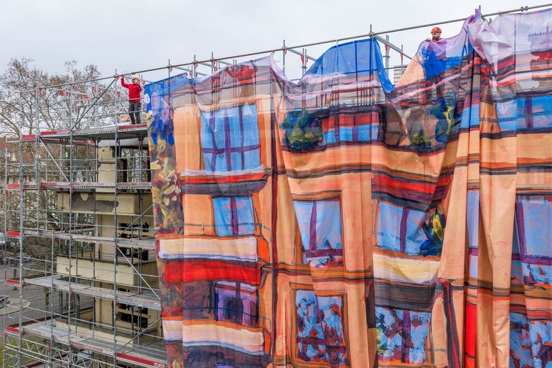 Ein mehrstöckiges Gebäude ist eingerüstet; mehrere Monteure befestigen oben eine grosse, farbig bedruckte Gitternetz-Blache am Baugerüst. Das Motiv zeigt eine gemalte Hausfassade und wirkt als Kunst am Bau – eine Grossformat-Reproduktion eines Acrylgemäldes von Michele Bordoni, umgesetzt als Grossformatdruck für die Gerüstverkleidung durch Richnerstutz.