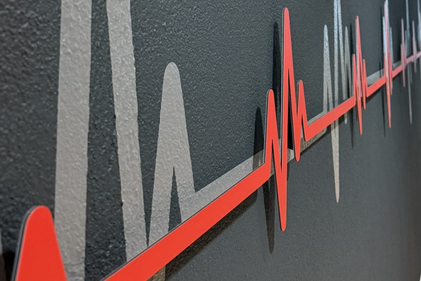Rote Herzschlag-Kurve aus Dibond mit 3D-Abstandshaltern vor grauer Folie an einer Wand.