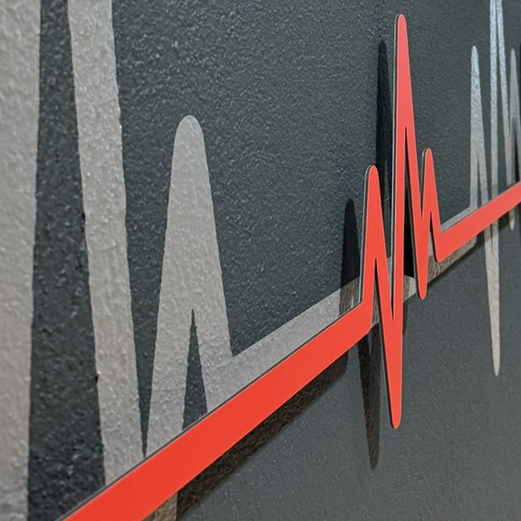Rote Herzschlag-Kurve aus Dibond mit 3D-Abstandshaltern vor grauer Folie an einer Wand.
