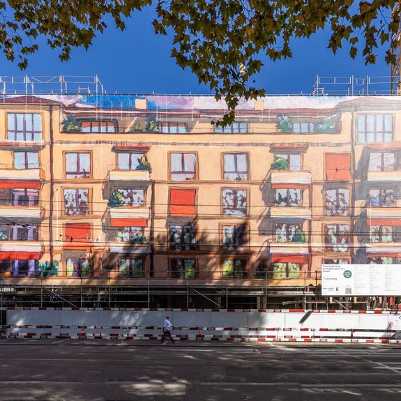 A close-up frontal view of an apartment building covered in scaffolding: A large scaffolding structure is draped with a printed mesh tarp that resembles facade art and traces the building’s facade as a colorful image. The large-scale scaffolding design was produced by Richnerstutz as a large-format print—based on an acrylic painting by Michele Bordoni, reproduced in high resolution by undknup AG, commissioned by Urs Ledermann.