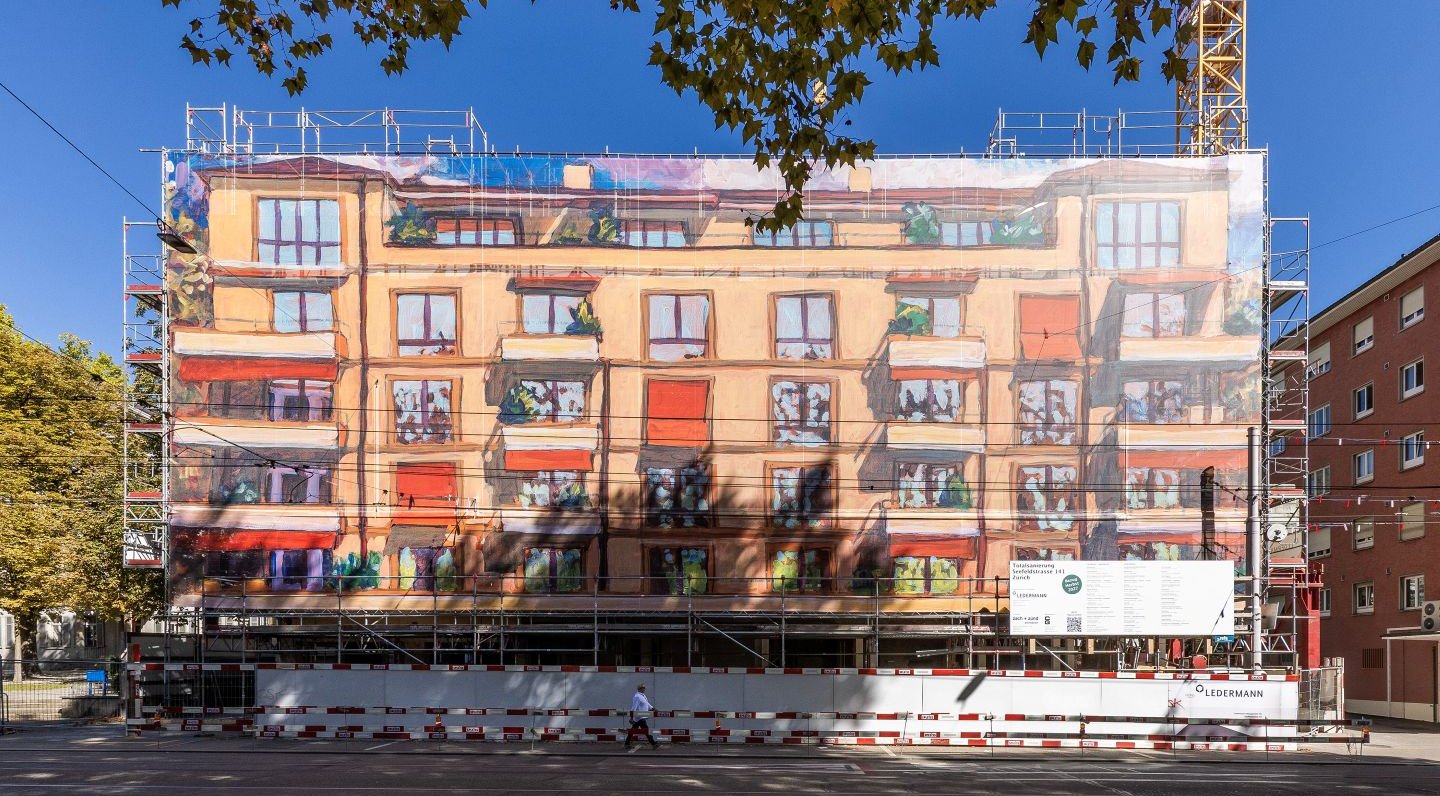 A close-up frontal view of an apartment building covered in scaffolding: A large scaffolding structure is draped with a printed mesh tarp that resembles facade art and traces the building’s facade as a colorful image. The large-scale scaffolding design was produced by Richnerstutz as a large-format print—based on an acrylic painting by Michele Bordoni, reproduced in high resolution by undknup AG, commissioned by Urs Ledermann.