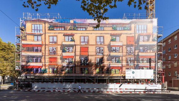 Gros plan frontal d'un immeuble d'habitation entouré d'échafaudages : un grand échafaudage est recouvert d'une bâche maillée imprimée qui fait office d'œuvre d'art de façade et reproduit la façade du bâtiment sous forme de motif coloré. Ce habillage de chantier grand format a été réalisé par Richnerstutz sous forme d’impression grand format, d’après une peinture acrylique de Michele Bordoni, reproduite en haute résolution par undknup AG, à la demande d’Urs Ledermann.
