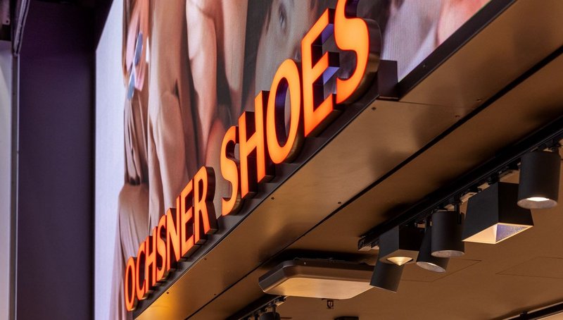 Eingangsbereich der Ochsner Shoes-Filiale mit grossem LED-Screen und davor platziertem Leuchtschriftzug, der eine starke Tiefenwirkung erzeugt.